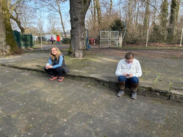 Zwei Personen sitzen auf einer Mauer, eine schaut auf ihrem Handy.