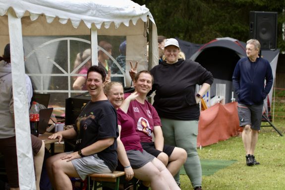 Fünf Personen sitzen in einem Garten, umgeben von Zelten und einem Pavillon.