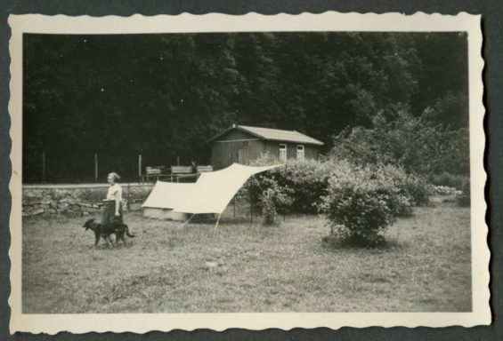 Ein Mann mit Hund vor einem Zelt und einer kleinen Hütte in der Natur.