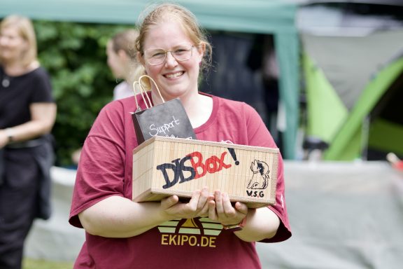 Frau mit rot-weißem Shirt hält eine Kiste mit dem Aufdruck „DB-Bock“.