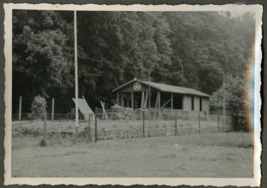 Ein älteres Haus in einer ländlichen Umgebung, umgeben von Bäumen und einem Zaun.