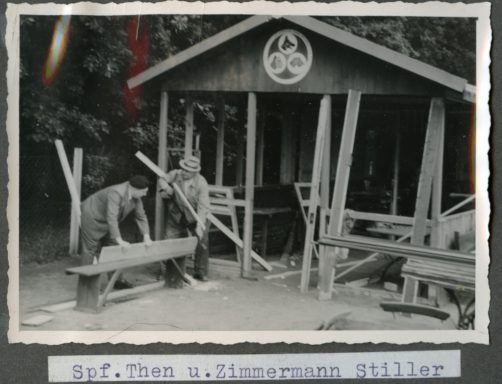 Zwei Tischler arbeiten an Holzarbeiten vor einem Bauwerk aus Holz im Freien.