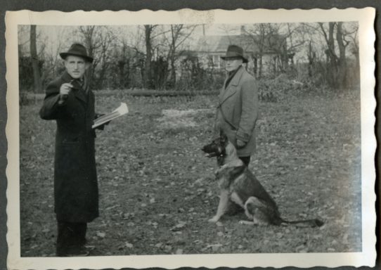 Zwei Männer in langen Mänteln stehen auf einem Feld, ein Hund sitzt neben ihnen.