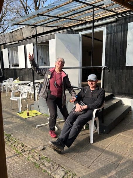 Zwei Personen, eine stehend und eine sitzend, vor einem schwarzen Gebäude mit Terrasse.