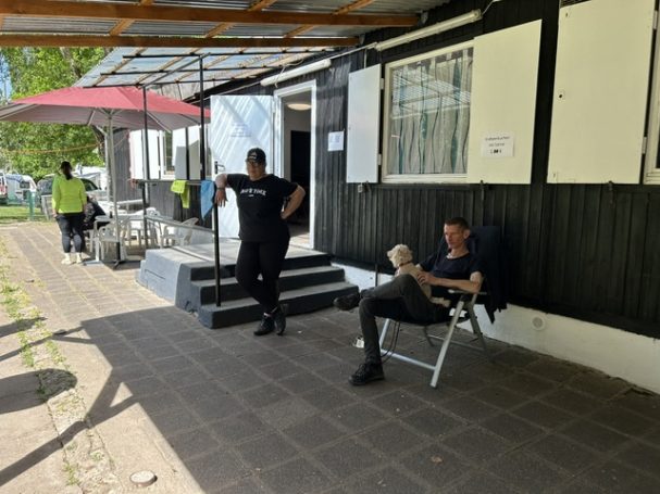 Zwei Männer stehen und sitzen vor einem Gebäude mit Sonnenschirm.