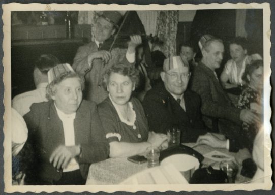 Eine Gruppe von Menschen in einem festlichen Raum, einige tragen Hüte, lachen und unterhalten sich.
