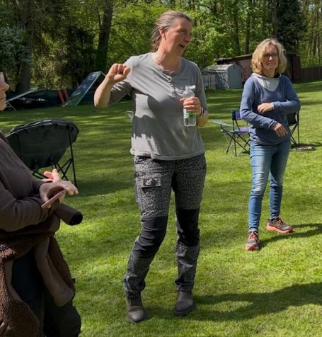 Drei Personen im Freien, eine gestikulierend mit Getränk, im Hintergrund Gartenstühle.