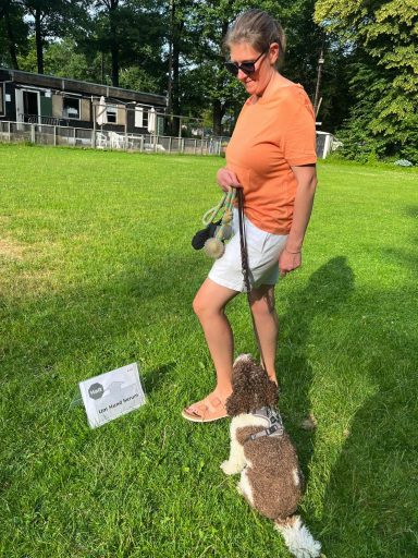 Frau mit Sonnenbrille steht auf einer Wiese neben einem Hund und einem Schild.