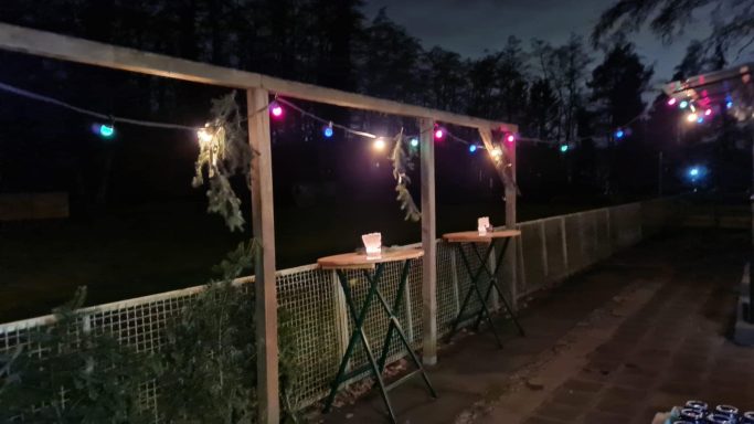 Markierte Holzpergola mit bunten Lichtern und zwei Tischchen in der Dämmerung.