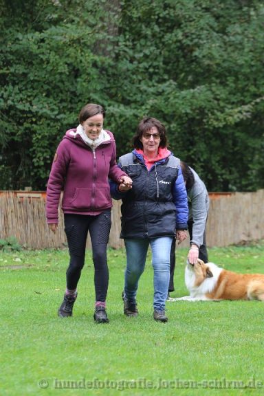 Zwei Frauen gehen auf einer Wiese, während ein Hund neben ihnen liegt.