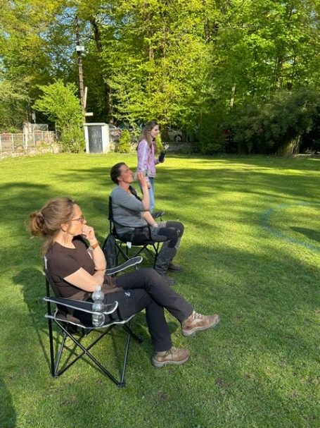 Drei Personen in Gartenstühlen stehen und sitzen auf dem Übungsplatz